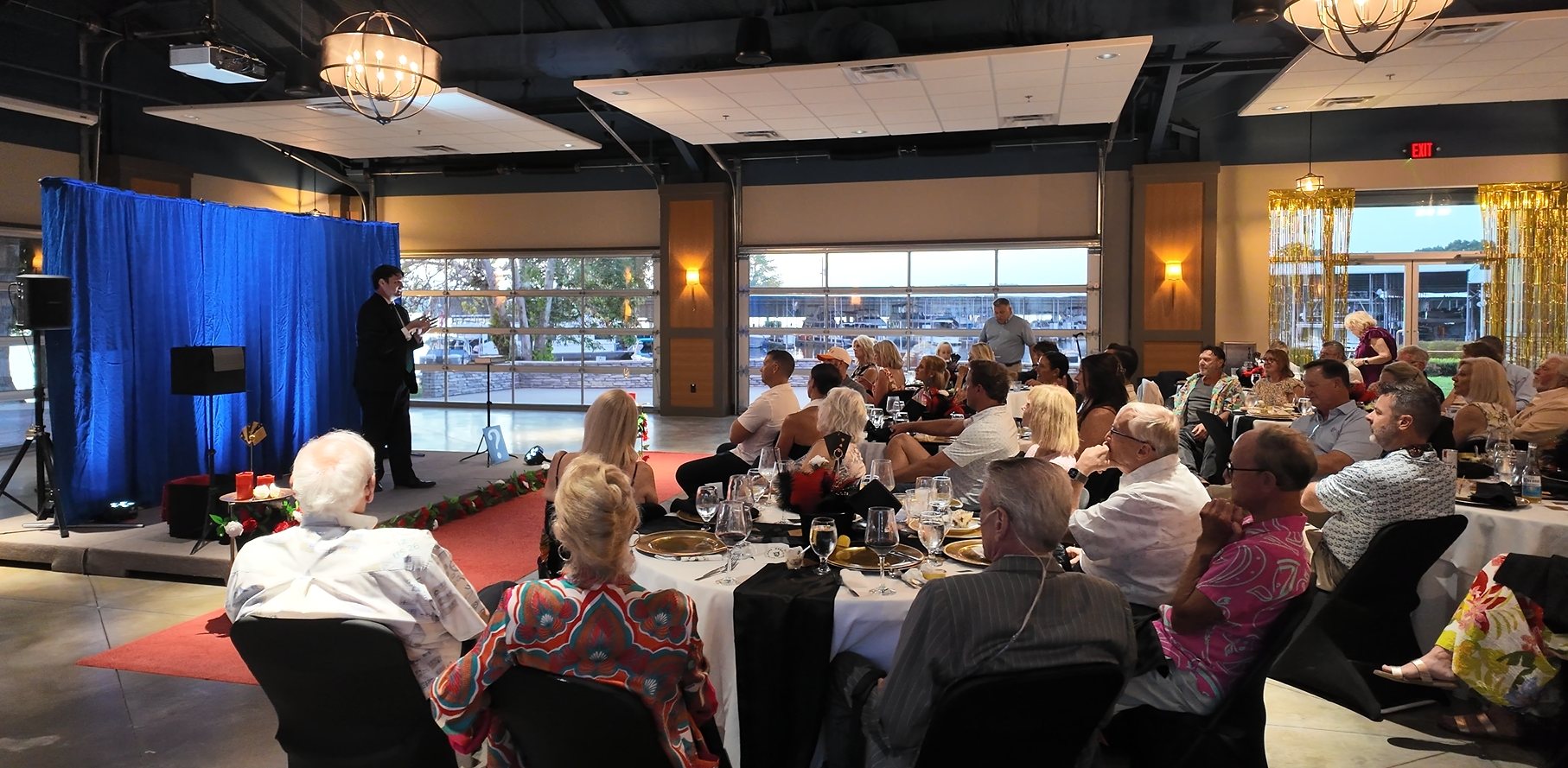 The Cedar Creek Yacht Club Supper Club in full, with members seated at round tables and Josh Weidner performing on the stage, the marina visible through the windows behind.